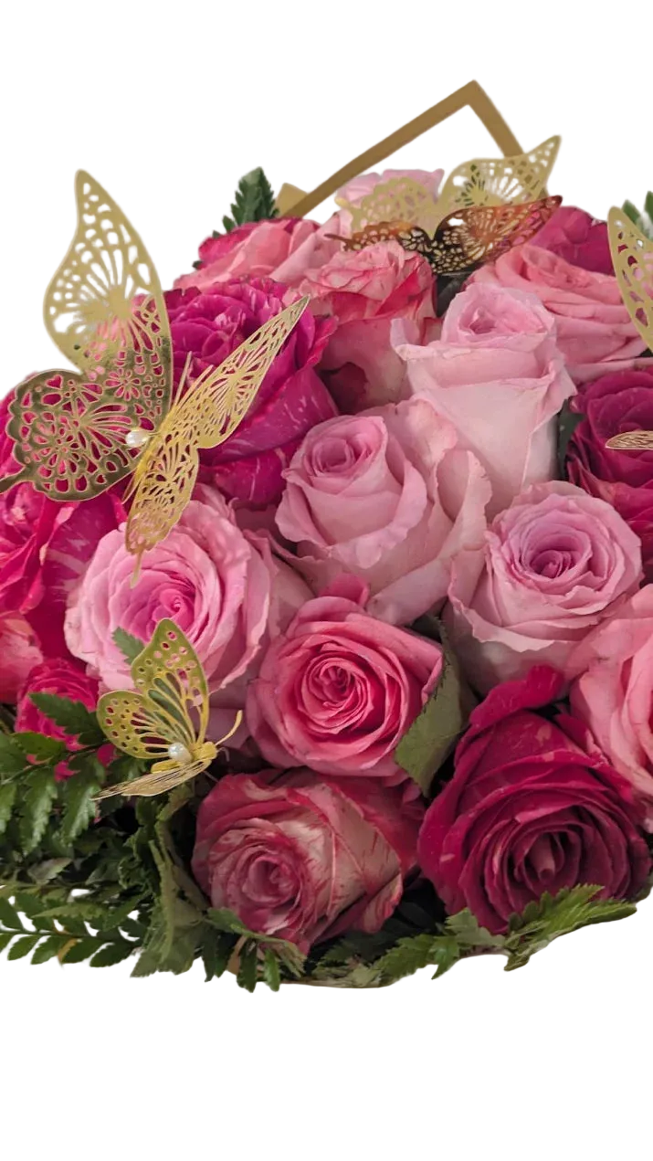 A bouquet of pink and magenta roses with green foliage, decorated with gold butterfly cutouts and a gold geometric accent, wrapped in white paper with a pink ribbon.
