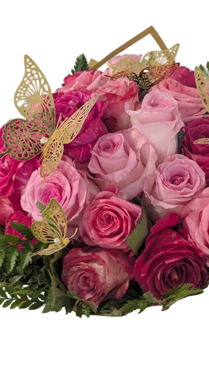 A bouquet of pink and magenta roses with green foliage, decorated with gold butterfly cutouts and a gold geometric accent, wrapped in white paper with a pink ribbon.