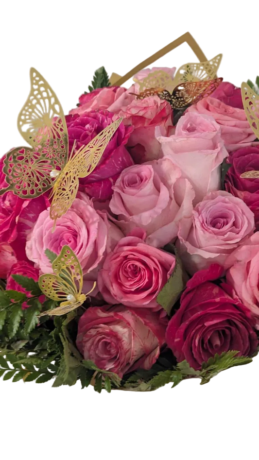 A bouquet of pink and magenta roses with green foliage, decorated with gold butterfly cutouts and a gold geometric accent, wrapped in white paper with a pink ribbon.